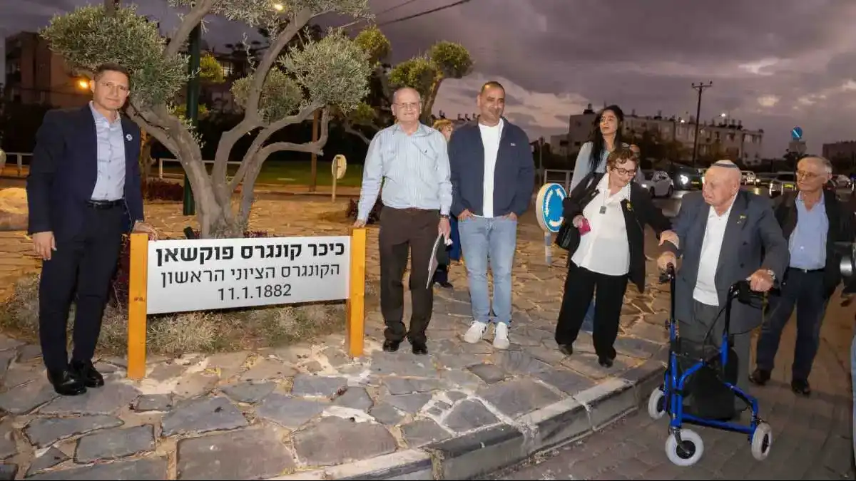 'טקס חנוכת כיכר פוקשאן בשכונה א' בבאר שבע. פרופ' ראובן איליה, ראש העיר רוביק דנילוביץ' ואישי ציבור ניצבים ליד השלט החדש של הכיכר, מוקפים במשתתפים וביוצאי הקהילה הרומנית.'