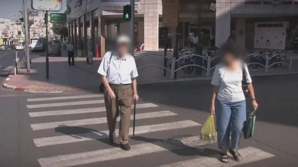 אזרח ותיק חוצה מעבר חצייה בעיר בישראל על רקע תנועה סואנת ותמרור בטיחות בדרכים